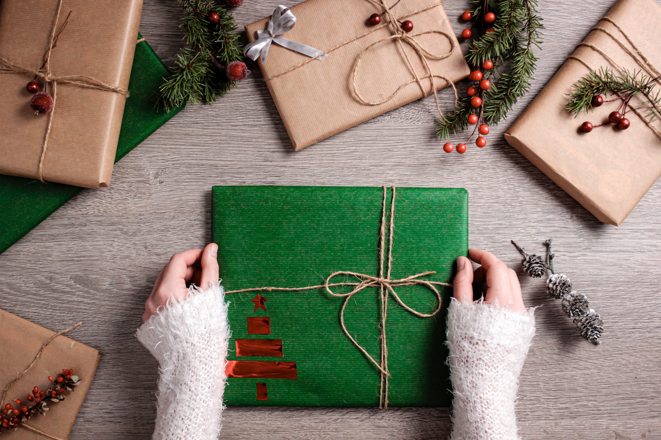 Deux mains en train d'emballer un cadeau avec du papier vert sur une table qui contient d'autres cadeaux emballés dans du papier brun.